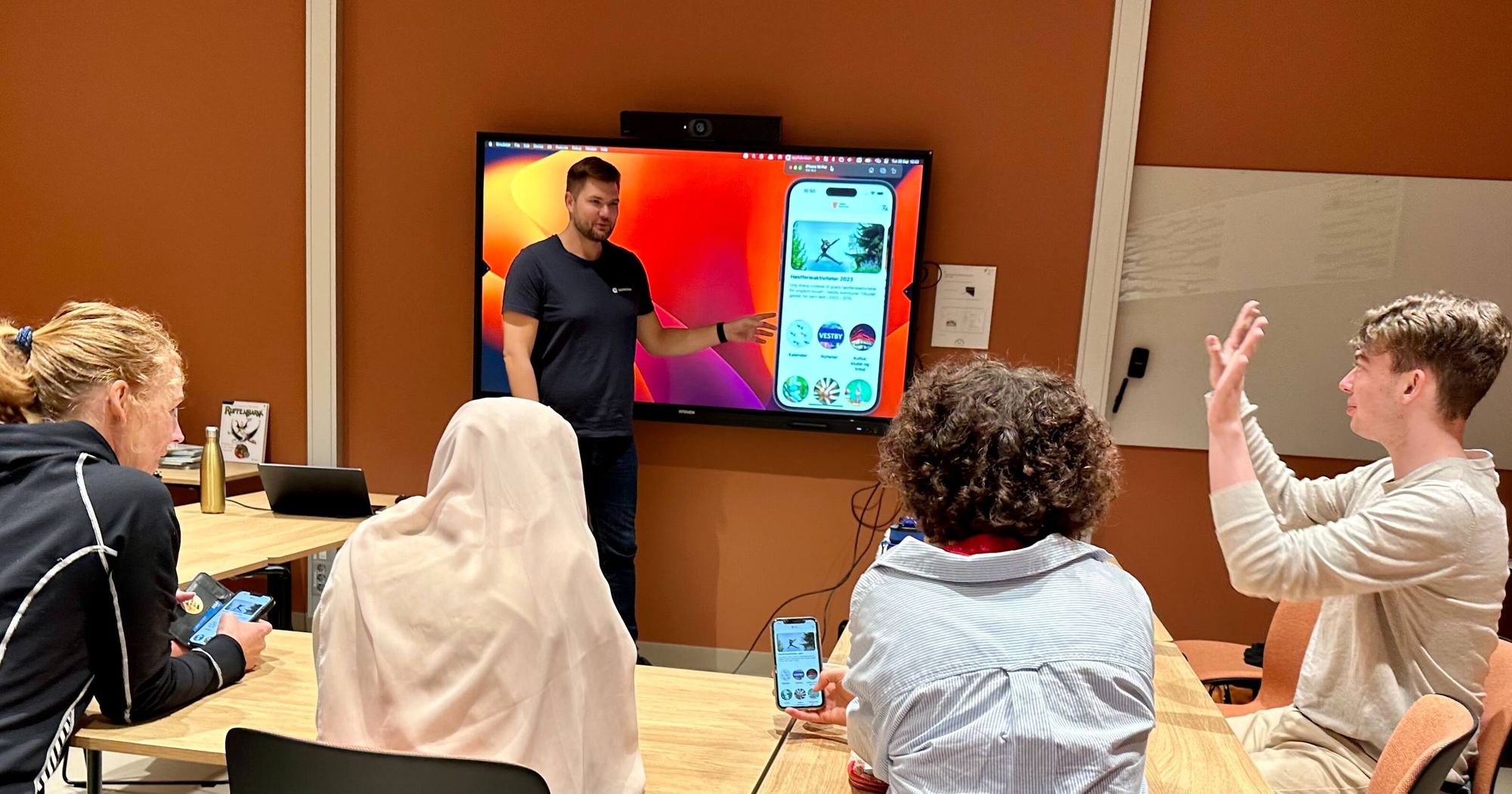 Fire personer rundt et bord ser på en person som presenterer en app på storskjerm. Foto.